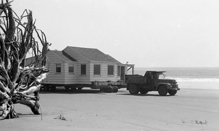 A white house has been placed on a trailer and hitched to the back of a truck that is towing it across a beach.