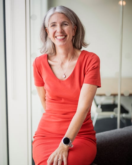 Rain Newton-Smith sits in her office on the back of a grey sofa. She wears a bright red dress with short sleeves, a scooped neck and a gold pendant necklace. She is smiling.