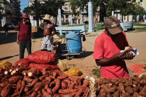 A pile of what appears to be sweet potatoes in a sunny square. A man stands next to them holding what appears to be a wrapped sweet potato. Two men stand behind him