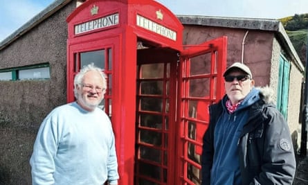 Two men pose for a photograph beside a classic ed phone box with its door open