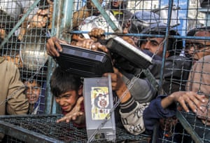 People holding plastic containers crowd around a small opening in a wire fence