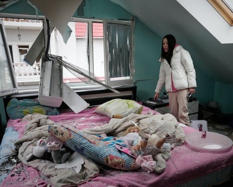 A woman standing in a room with a damaged ceiling looks at debris on a bed.