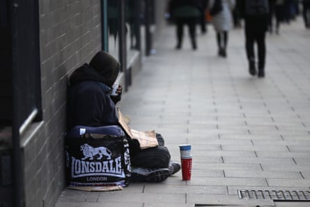 A man wearing a hooded top and begging for small change sits on a pavement with his back against a dark brick wall. He has bags around him, a stack of cardboard cups in front and is holding a cardboard sign.