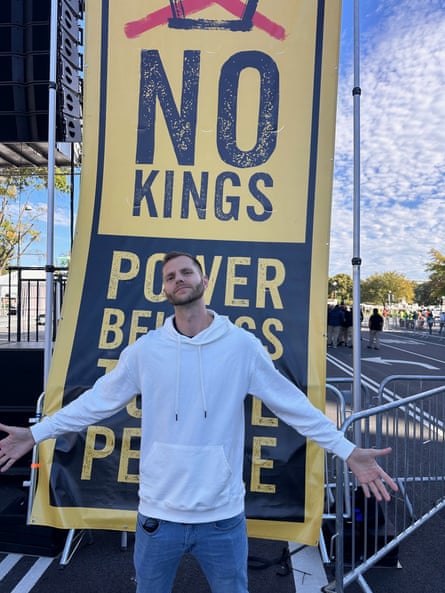 man stands outside at protest with arms out in front of large sign saying ‘no kings / power belongs to the people’