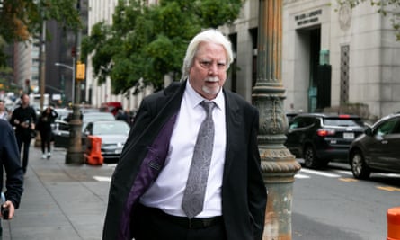 A tall white man with shaggy bright white hair looks angrily at the camera, walking in a dark suit and dark trenchcoat down a city street