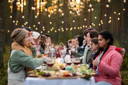 In woods lit by fairy lights, people in winter garb sit eating at a table loaded with food