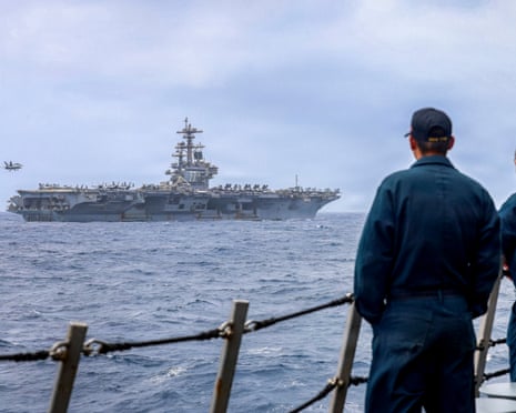 A sailor in dark uniform watches an aircraft carrier with jets on deck from another vessel