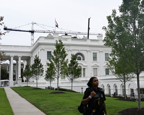 A Secret Service officer stands on the White House lawn, with a crane visible above the roof