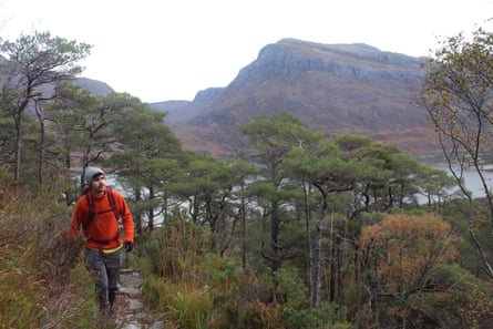 A young man with moustache in hiking gear walking through a pinewood with a loch and mountain in the background