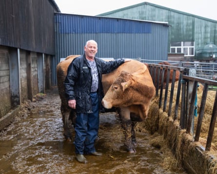 A farmer wearing overalls stands next to a brown cow stroking her back. There is a barn behind.