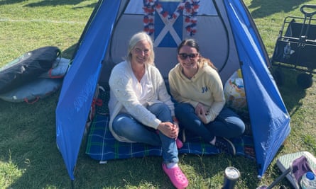 Christine, 63, and her daughter Lorna, 40: they are sitting in the entrance of a small blue tent on a tartan rug, with a Scottish flag and a garland of red, white and blue flowers seen hanging above the tent’s opening. Christine has grey hair and wears a white shirt and cardigan, blue jeans and bright pink Croc rubber clogs, while her daughter has long dark hair in a pony-tail and wears sunglasses, a pale yellow hoodie, and blue jeans and trainers. They look happy in the sunshine.