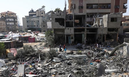 People walk among debris in the Nuseirat camp in the aftermath of Israeli strikes; ruined buildings are seen in the background
