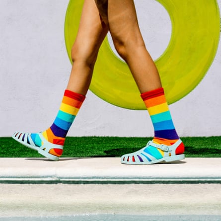 A bright summer scene with legs in colourful rainbow socks and sandals walking by a pool, next to a yellow inflatable ring. Green grass adds to the vibe.
