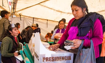 A woman in a bright pink dress places a ballot into a box marked "presidencia".