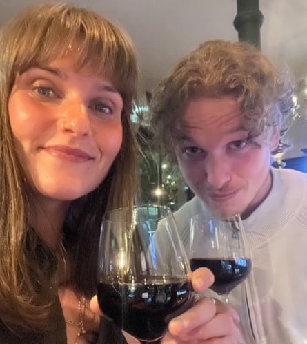 A smiling young woman with long blond hair and a fringe, and a young man with curly brown hair, both holding glasses of red wine