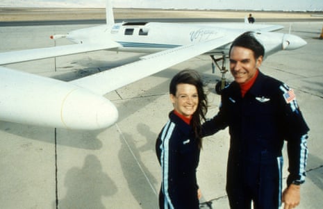 Two young-middle-aged white people in dark blue jumpsuits with white piping smiling broadly in obvious wind in front of a low white plane.