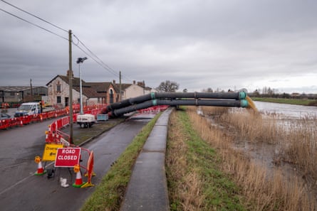 huge black pipes across the footpath are pumping water into the river. Brick and stone village buildings and a large tin-roofed barn are in the background; the landscape is flat and the sky is grey.
