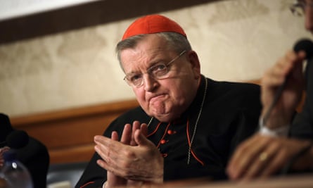 Older white man with glasses, red skull cap, black outfit with red piping, claps and purses lips.