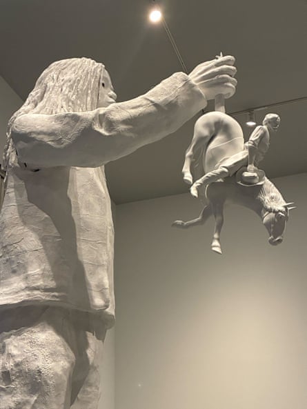 White sculpture of large Black figure holding tiny horse and rider by the tail.