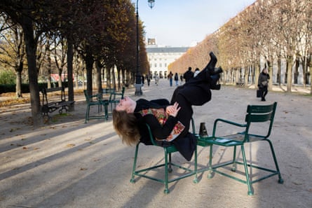A young woman sits on a chair in a formal French city garden kicking her legs up 