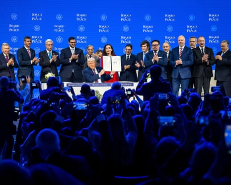 An old man looking like an orange waxwork holds up paper with a signature while people stand around him applauding