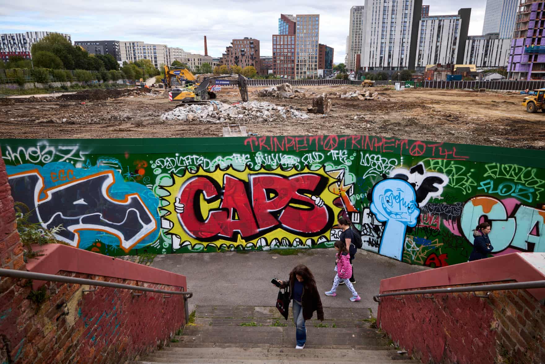 People descend steps past graffiti near Ancoats, with cleared demolition land and city towers behind
