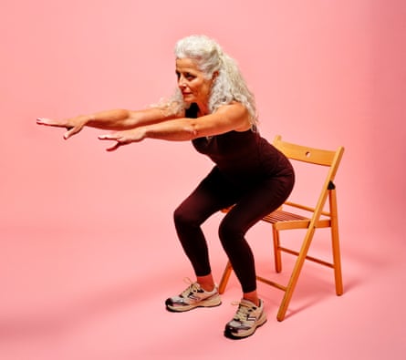 A woman with long white curly hair wearing a black vest and leggings, doing a squat in front of a folding wooden chair