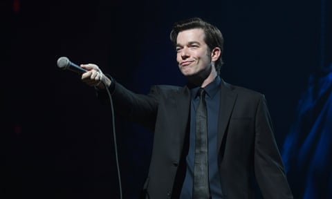 A man in a suit and tie holds a microphone up to the air with a smug grin on his face