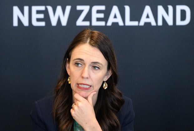 Jacinda Ardern looking thoughtful in front of a backdrop with the words New Zealand on it
