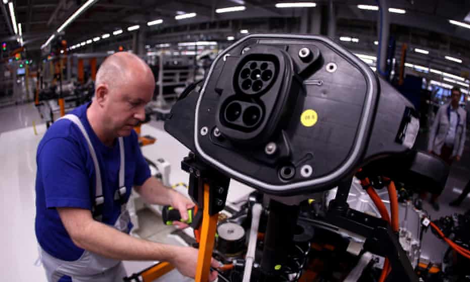 Photograph of an assembly line worker working on an electric car, with car's charging socket prominent in the camera's view