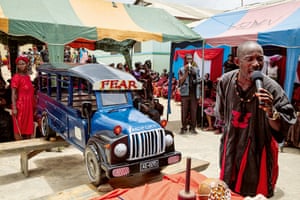 A large truck-shaped coffin in blue and black resting on a platform with a man speaking into a microphone and colourful tents erected nearby
