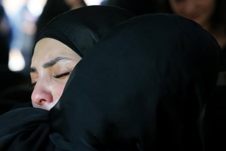 The pained face of a woman wearing a black hijab is visible over the shoulder of another woman in a black hijab who is facing away from the camera, hugging the other woman.