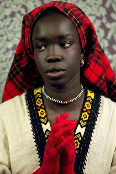 A woman wears a red tartan headscarf and embroidered clothing, hands clasped in red gloves