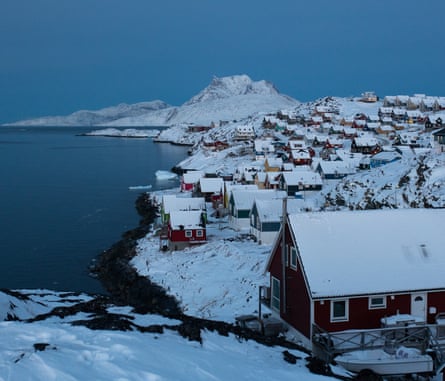 A photograph of houses with snow covered roofs lining the coast of a sea or lake, with hills behind also covered in snow