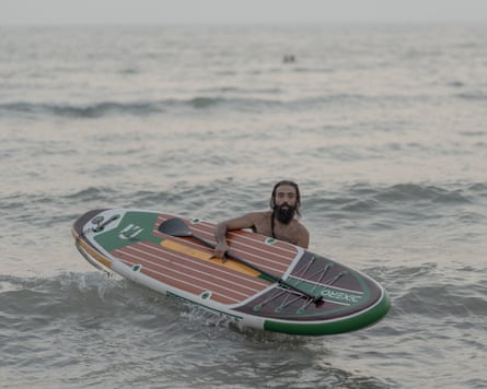 Yashar stands in the sea looking at camera while holding board and paddle