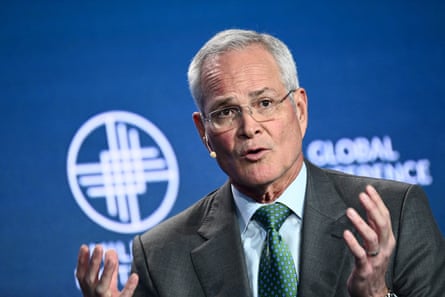 A man wearing glasses, a suit, and tie gestures with his hands while speaking at a conference