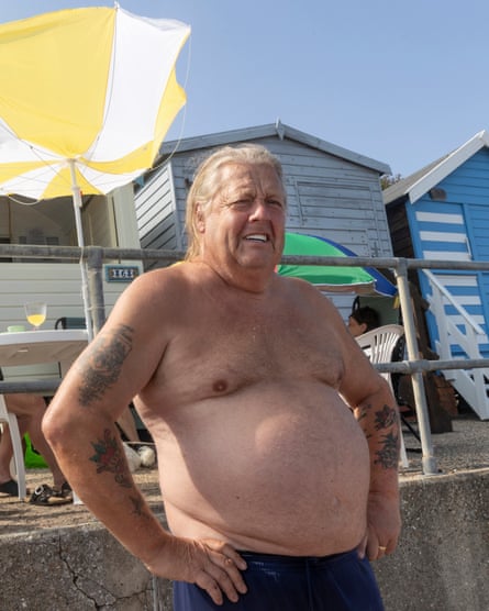 A large man with his top off stands in front of beach huts
