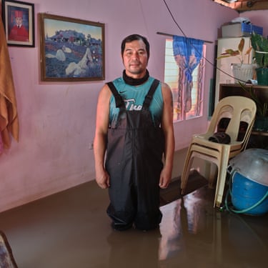 A man in overalls stands inside a house with pink walls and shin-deep in flood water
