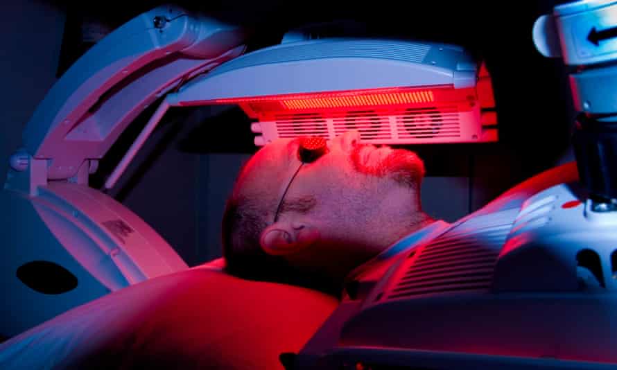 The face, with goggles over his eyes, and covered torso of a male patient lying on a bed with an LED red light machine hovering over his face
