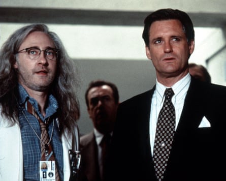 Two men standing, one in a white lab coat and the other in a suit, with others behind them