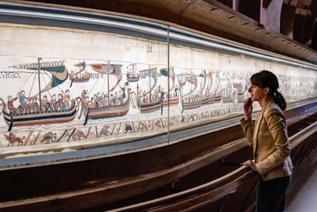 A woman stands by the tapestry which is in an illuminated display case; she is viewing a section showing a fleet of ships sailing from France, and the room is darkened.