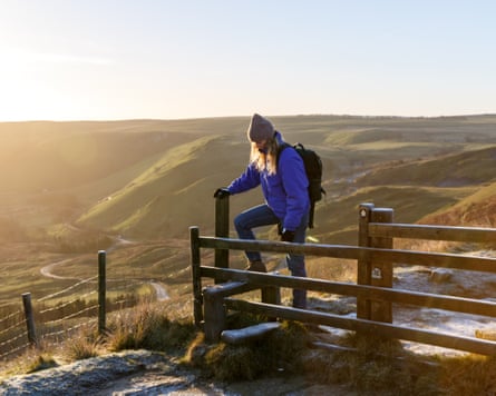 A young woman walks over a stile at sunrise in the Peak District national park, England. Taken on the Mam Tor ridge at sunrise in the Peak District national park, Edale, England. Woman is a young hiker wearing hiking clothes and a backpack.