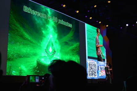 A view from the audience of a bright green screen in a darkened auditorium and a man standing in front of it at the far end.