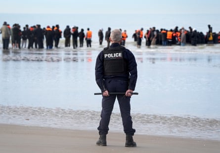 A police officer with back to camera looks on as people stand in shallow water trying to board a large rib-type boat