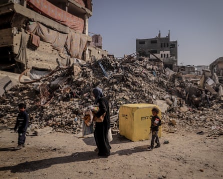 A woman and two children pass a yellow concrete block near a pile of rubble