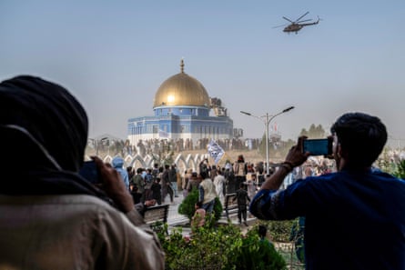 A helicopter flies over a mosque with gold dome on a hilltop where men are standing; two in the foreground are holding up phones to take photographs