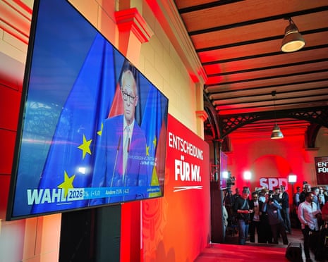 A screen shows Chancellor Friedrich Merz speaking, beside an SPD election party banner in Mecklenburg-Western Pomerania