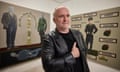 Ignacy Czwartos wears a leather jacket and black t-shirt. He stands with his hand on his heart in front of two pieces of his work for Polonia Uncensored.