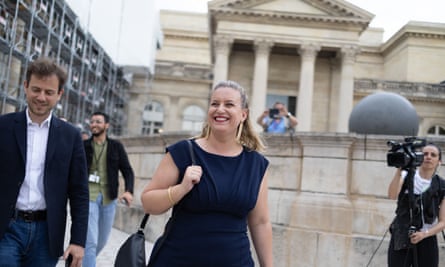 Mathilde Panot arrives at the National Assembly, Paris, 9 Jul 2024: she is seen in front of a pale stone building with pillars and classical-style entrance, with a photographer to her side; she is in her mid-30s and has long blond hair tied back, and wears a sleeveless black dress and dangling earrings. She is smiling.