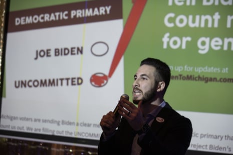 A man speaks into a microphone in front of a projection showing a ballot with a check next to 'uncommitted' and an empty cirle next to "Joe Biden'
